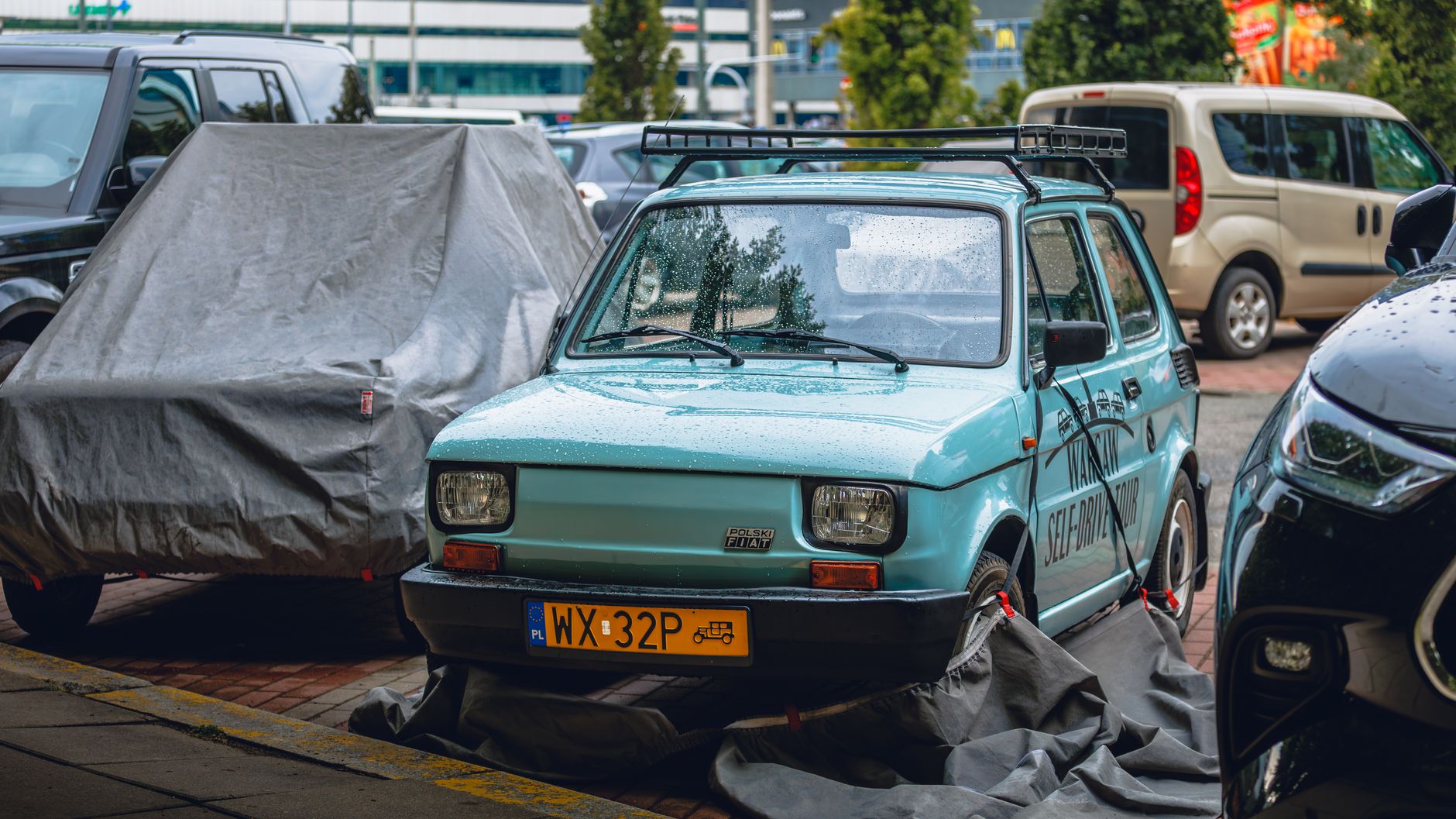 Fiat 126p zaparkowany w deszczu między nowoczesnymi samochodami